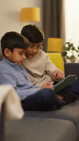 Vertical-Video-Of-Two-Young-Boys-Sitting-On-Sofa-At-Home-Playing-Games-Or-Streaming-Onto-Digital-Tablet-Together-1